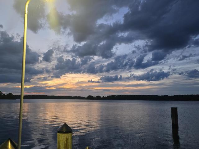 A wooden dock with posts extends over a calm lake at sunset, under a cloudy sky. A tall lamp post is lit on the left, reflecting on the water’s surface.