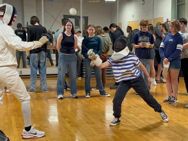 Two people fence in a gym while a group of onlookers watch; one fencer is lunging, and the other stands ready.