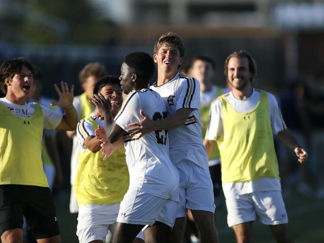 Soccer players in white uniforms celebrate together on the field, with teammates in yellow pinnies joining in and smiling in the background.