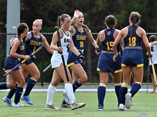 A field hockey team in navy and yellow uniforms celebrates on the field as two players in white walk away, with trees and a fence in the background.