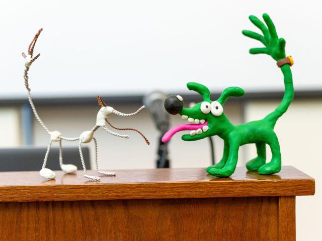 Two dog sculptures, one made of wire and white beads, the other made of green clay with exaggerated features, are displayed on a wooden surface.