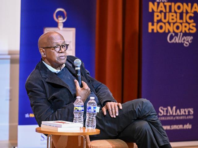 A man sits on stage holding a microphone, with two water bottles and a book on a table beside him. A "National Public Honors College" banner is in the background.