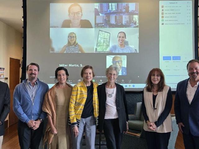 Eight people stand in a row indoors, with a large screen behind them showing a video call with five additional people.