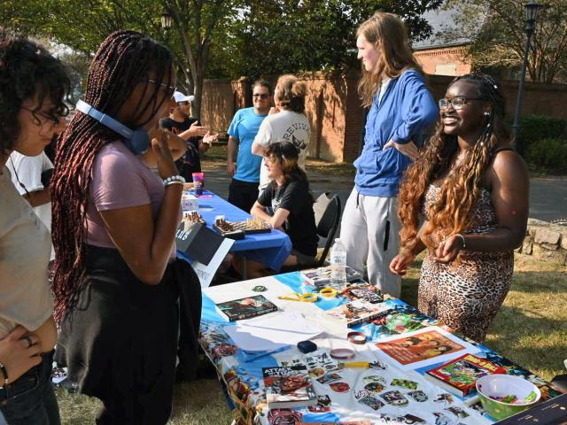People interact at an outdoor table covered with colorful artwork and crafts during a community or school event on a sunny day.