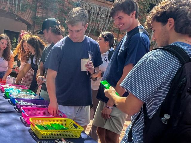 A group of young adults browse colorful baskets of items on a table at an outdoor event. One person holds a drink, another examines a green object.