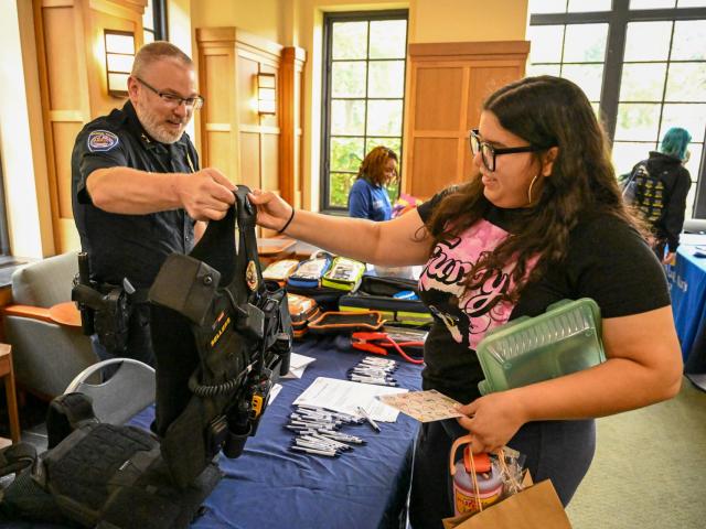A police officer shows a tactical vest to a woman at an indoor event table with informational materials and giveaway items.