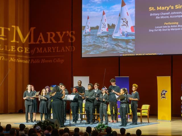 A choir performs on stage at St. Mary’s College of Maryland, with a projected presentation and audience seated in the foreground.