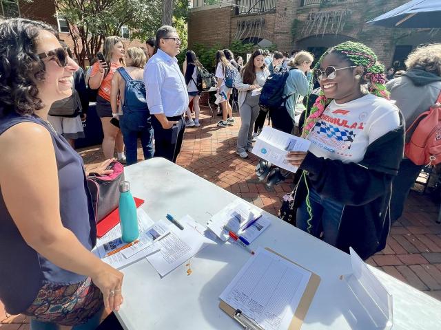 Two women stand at a table with papers and pens, talking and smiling, while other people gather at an outdoor event with booths in the background.