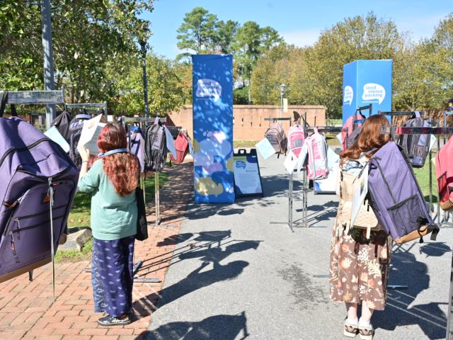 Two people stand among rows of large backpacks displayed outdoors on metal racks, with informational signs and trees in the background.