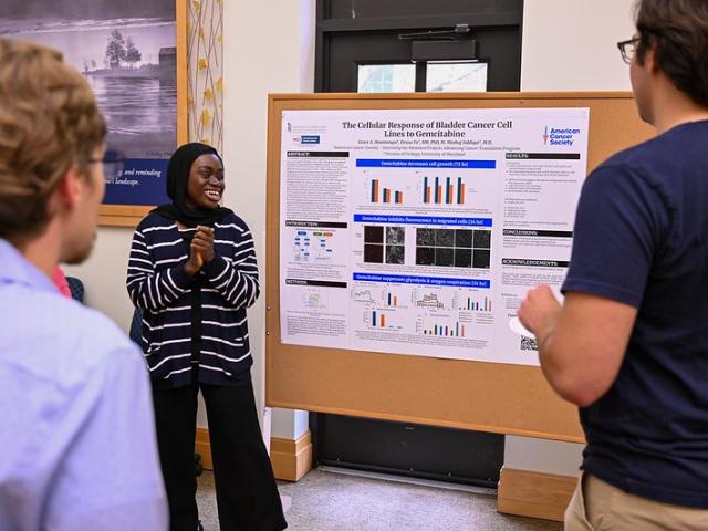 A woman presents scientific research on cancer cell response to three people in front of a poster display in a bright room.