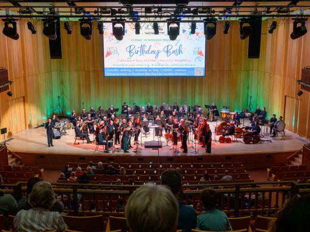 A symphony orchestra performs on a well-lit stage in a concert hall, with musicians seated and some standing; audience members watch from the seats.