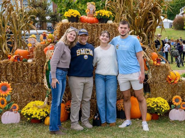 Four people pose in front of a fall-themed display with hay bales, pumpkins, sunflowers, and autumn decorations outdoors.