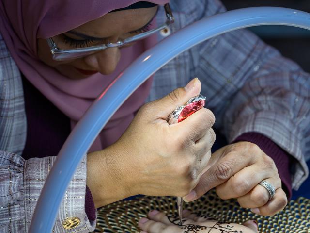 A person in a hijab applies intricate henna designs on another person's hand under a bright ring light.