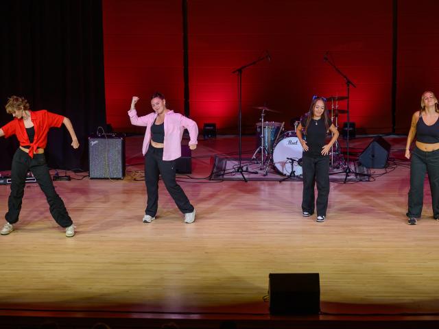 Four people perform a dance routine on stage in front of musical instruments and microphones, with red lighting in the background.