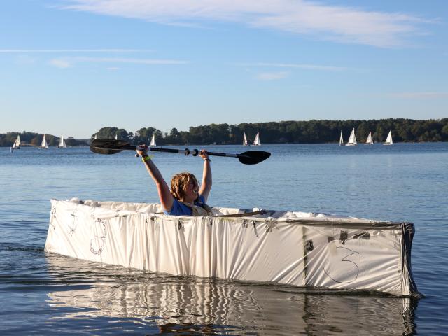 A person sits in a homemade cardboard boat on a lake, raising a paddle overhead. Sailboats are visible in the background under a clear sky.