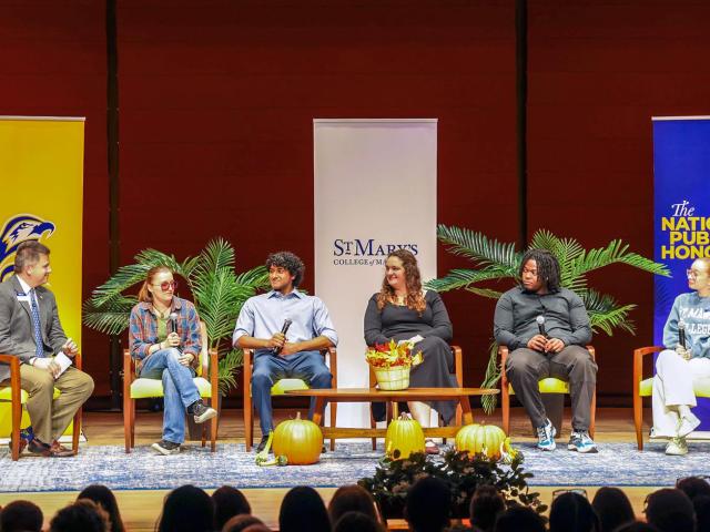 Six people sit on stage in chairs during a panel discussion, with banners, plants, and pumpkins as decoration and an audience in the foreground.