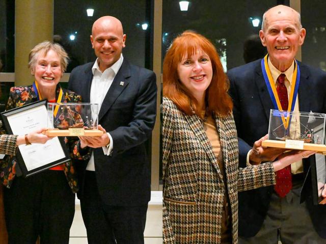 Four people pose indoors; on the left, a woman holds an award and another holds a certificate. On the right, the same woman presents the award to an older man with a medal.