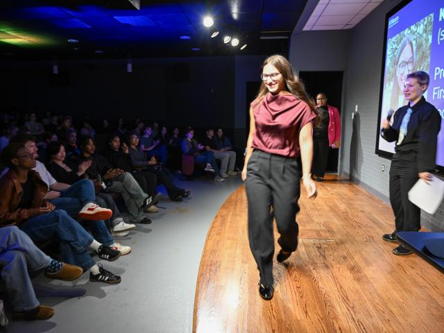 A person walks across a stage in front of an audience, while two people stand by a screen displaying a professional profile and photo.