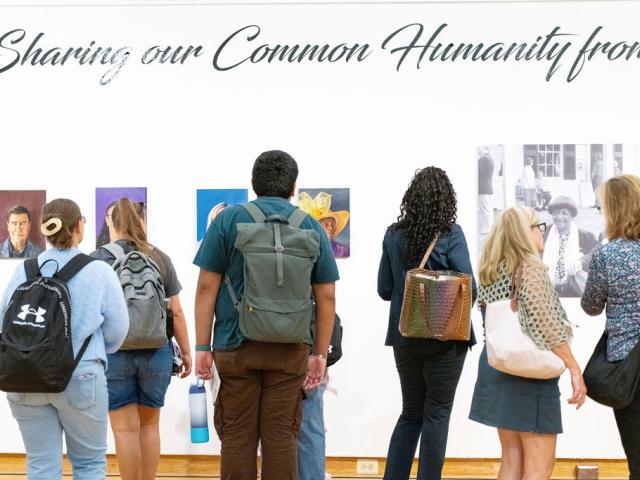 A group of people with backpacks and bags view artwork on a gallery wall beneath a sign that reads "Sharing our Common Humanity.