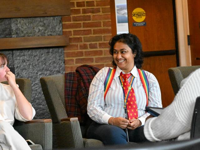 Three people sit in chairs having a conversation; one person in the center wears a striped shirt, rainbow suspenders, and a red tie, smiling at another person.
