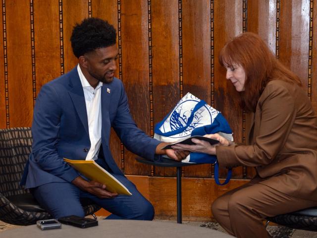 A man in a blue suit and a woman in a brown suit sit in chairs by a small table, looking at a tablet together. Blue-and-white tote bag and documents are on the table.