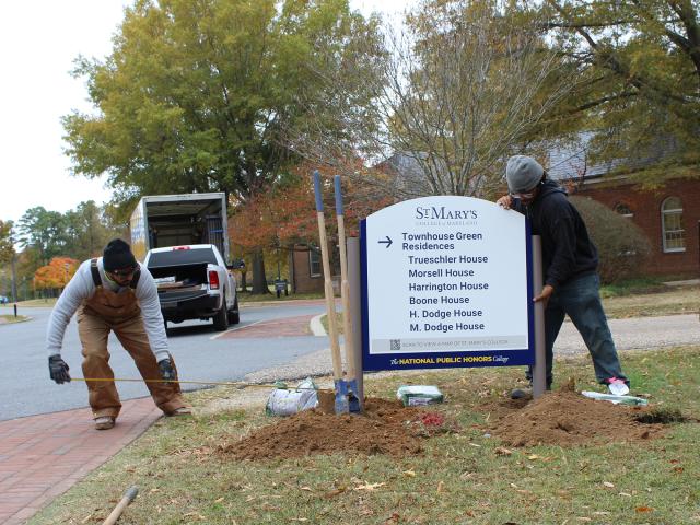 Two workers install a St. Mary's College directional sign listing campus residences, with soil and tools on the ground nearby. A pickup truck is parked in the background.