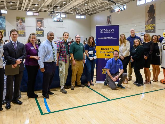 A group of people stands in a gymnasium next to a “St. Mary’s Career & Internship Fair” banner, with tables and a mascot visible in the background.