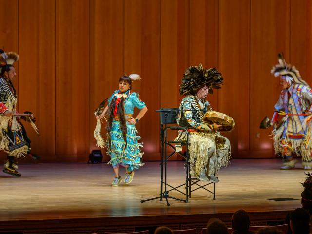 Four people in traditional Native American regalia perform on a wooden stage; one seated person plays a drum while three others dance in the background.