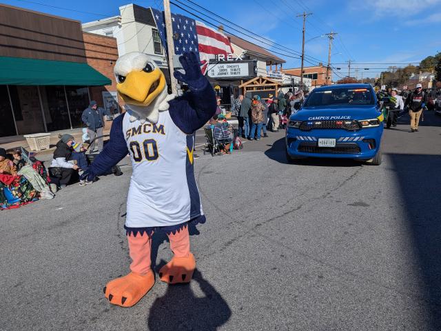 A person in an eagle mascot costume wearing an SMCM jersey walks in a street parade next to a blue campus police car, with spectators on both sides.