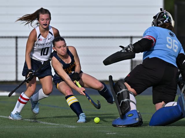 A field hockey player in blue attempts to hit the ball past a goalie in blue gear, while another player in white follows closely behind.