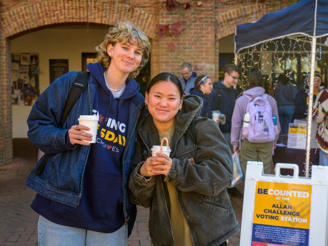 Two people stand outside near a voting station sign, holding drinks and smiling at the camera; other people can be seen in the background.