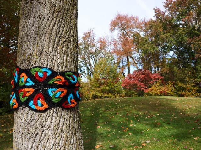 A tree trunk wrapped with a colorful crocheted yarn pattern stands in a grassy park with autumn foliage in the background.
