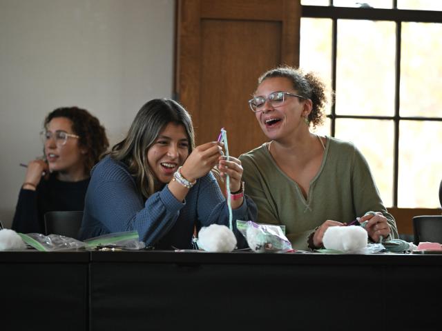 Two women sit at a table, smiling and laughing while holding craft supplies. Other people are seated nearby, and the table has bags of materials and cotton balls.