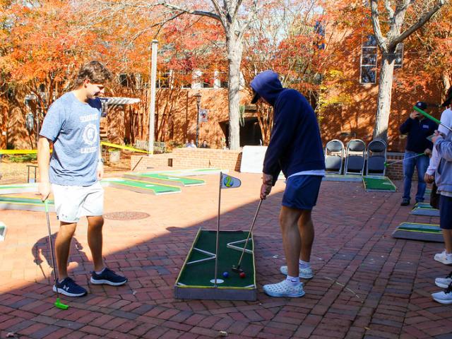 Several people play mini-golf on a brick courtyard surrounded by autumn trees and a brick building under sunny weather.