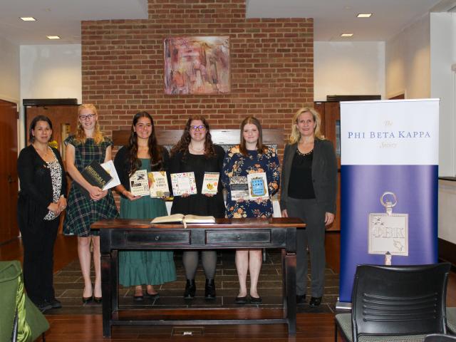 Six women stand indoors behind a desk with books, next to a "Phi Beta Kappa Society" banner. They are dressed formally and looking at the camera.