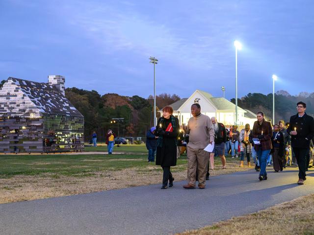 A group of people walk on a paved path in a park at dusk, with a metallic house-shaped structure and pavilion visible in the background.