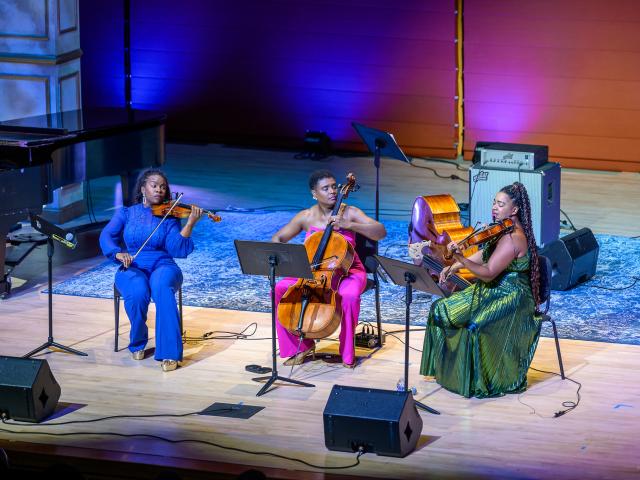 Three musicians perform on stage, playing violin, cello, and viola. They are seated with music stands on a lit wooden floor with a rug and stage lights in the background.