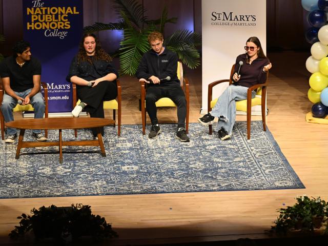 Four people sit on stage for a panel discussion at St. Mary's College of Maryland, with banners, plants, and balloons in the background.