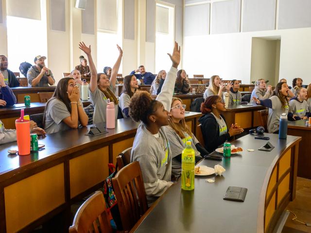 Students in a classroom sit at curved desks, some raising their hands excitedly while looking toward the front, with drinks and notebooks on the desks.