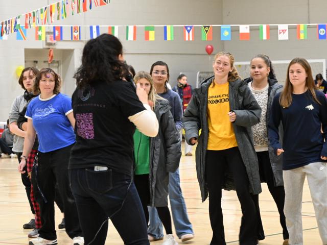 A group of young people standing in a gymnasium, listening to a woman speaking, with international flags hanging above them.