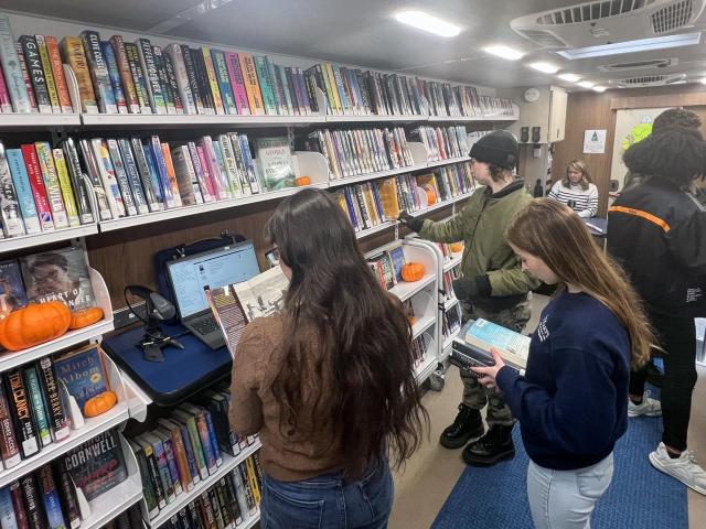Four people browse and read books inside a mobile library filled with colorful book spines and several small pumpkins on the shelves.