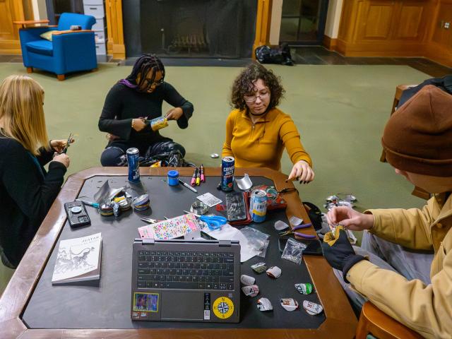Four people sit around a table working on arts and crafts with various supplies, drinks, a laptop, and books in a lounge or common room setting.