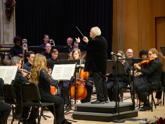 A conductor leads an orchestra of musicians playing string and brass instruments on a stage during a performance.