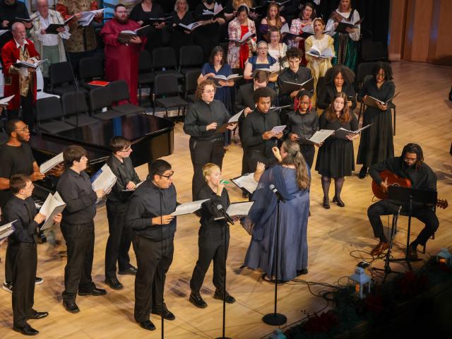 A choir and musicians perform on a stage, with a conductor leading them. Singers hold sheet music and a guitarist is seated to the right. Audience members are visible behind them.
