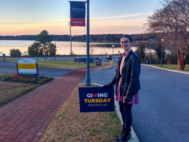 A person stands on a sidewalk holding a "Giving Tuesday #SMCMGIVES" sign near a campus entrance with a river and trees in the background at sunset.