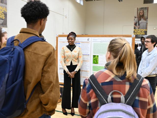 A student stands in front of a science poster presentation, speaking to a small group of people in a gymnasium setting.