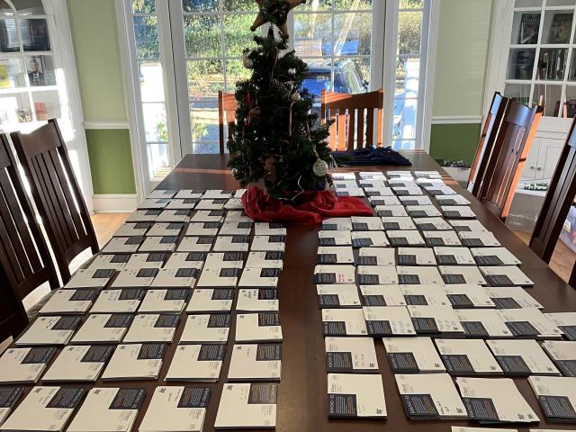A dining table covered with rows of cards or envelopes, with a decorated Christmas tree centerpiece and sunlight streaming in through glass doors.