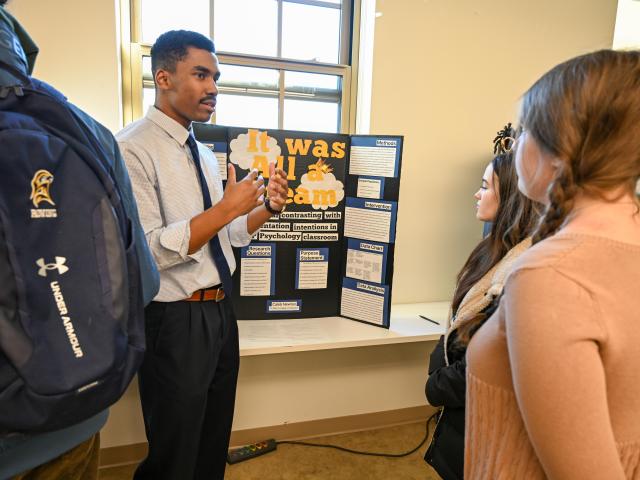 A man presents a project titled "It was All a Dream" on a trifold board to three people in a classroom setting.