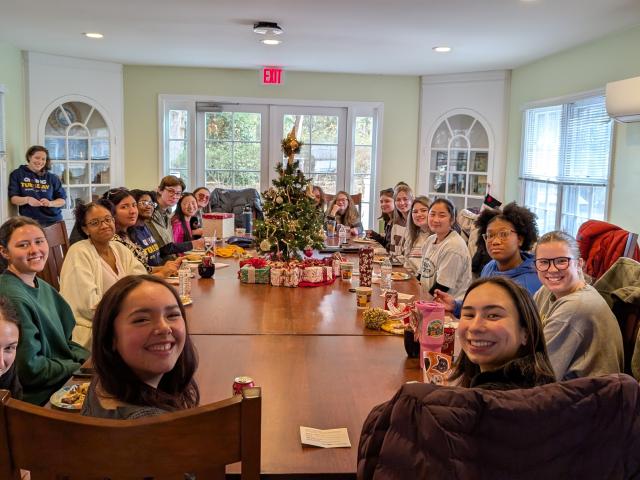 A group of people sit around a large table with food, drinks, and a small decorated Christmas tree in the center, gathered in a bright, cozy room.