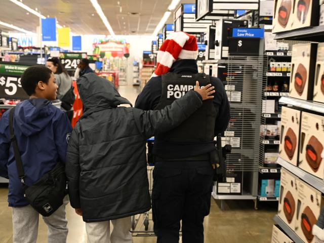 Two boys walk with a campus police officer wearing a Santa hat in a store aisle lined with boxed products.
